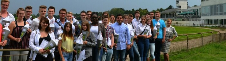 Neue Auszubildende der ATLANTIC Hotels vor der Galopprennbahn in Bremen Gruppe von Menschen steht auf einer grünen Wiese vor einem modernen Gebäude bei bewölktem Himmel.