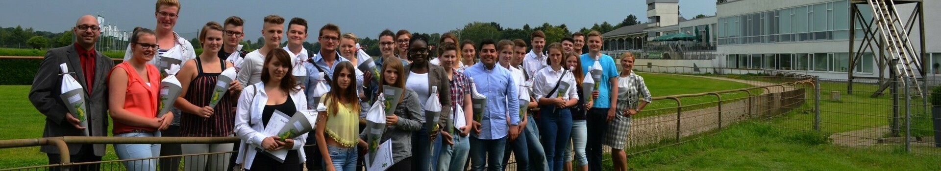 Neue Auszubildende der ATLANTIC Hotels vor der Galopprennbahn in Bremen Group of people standing on a green meadow in front of a modern building under a cloudy sky.