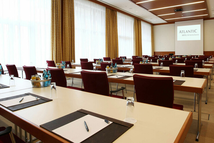 Heller Konferenzraum mit Reihen von Tischen, roten Stühlen, Schreibmaterial und Getränken, Projektionsfläche im Hintergrund.