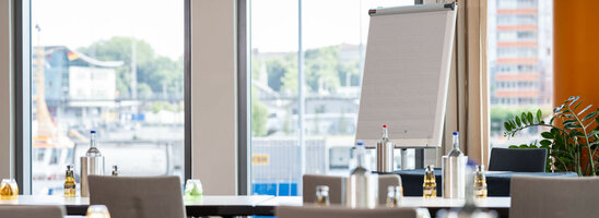 Bright conference room in the ATLANTIC Hotel Kiel with flipchart, tables and chairs, view of the harbor through large windows.
