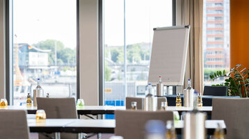 Heller Konferenzraum im ATLANTIC Hotel Kiel mit Flipchart, Tischen und Stühlen, Blick auf den Hafen durch große Fenster.