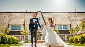 Brautpaar vor dem reetgedeckten Severin*s Resort & Spa auf Sylt, umgeben von gepflegten Gärten und Sonnenschein.