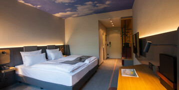 Modernes Hotelzimmer mit Doppelbett, Schreibtisch, TV und Wolkenmotiv an der Decke im ATLANTIC Hotel Airport Bremen.