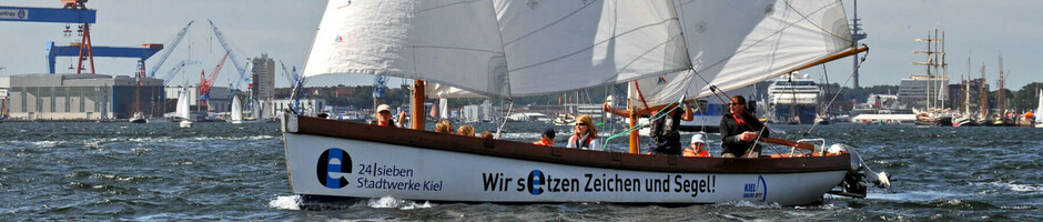 Segelboot mit drei Segeln auf der Kieler Förde, im Hintergrund Werft und weitere Boote.