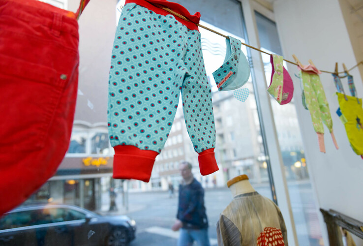ATLANTIC Hotel Sail City | Upcycling-Offensive "GRÜNER FADEN" Bunte Kinderkleidung hängt in einem Schaufenster in Bremerhaven, mit Blick auf die Straße im Hintergrund.