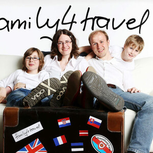 Familie sitzt lächelnd auf einem Sofa hinter einem Koffer mit Länderflaggen-Aufklebern.