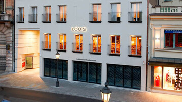 Außenansicht des LOUIS Hotels in München bei Dämmerung, mit beleuchteten Fenstern und angrenzenden Geschäften.