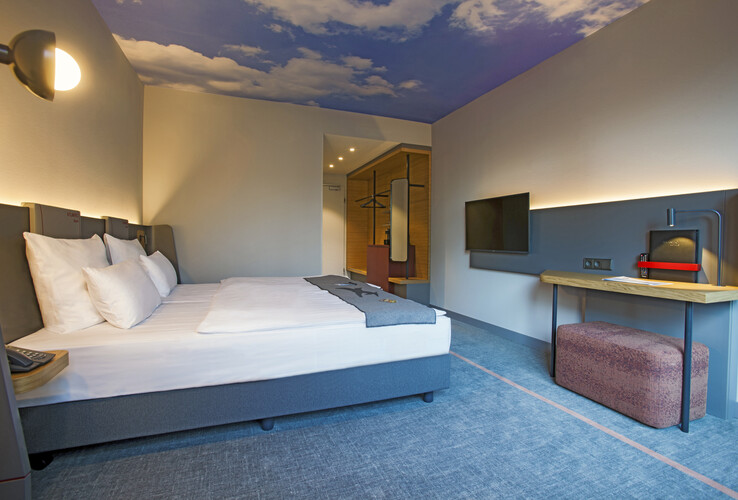 ATLANTIC Hotel Airport – Business Zimmer  Modernes Hotelzimmer mit Doppelbett, Schreibtisch, Flachbild-TV und Himmelmotiv an der Decke im ATLANTIC Hotel Airport.