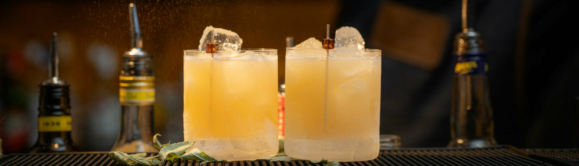 Zwei Cocktails mit Eiswürfeln auf einer Bar, ein Barkeeper verfeinert sie mit Zitrusnote.