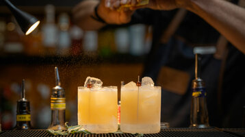 Zwei Cocktails mit Eiswürfeln auf einer Bar, ein Barkeeper verfeinert sie mit Zitrusnote.