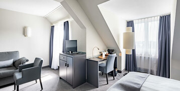 Helles Hotelzimmer mit Doppelbett, Schreibtisch, Fernseher, Sofa und großen Fenstern mit blauen Vorhängen.