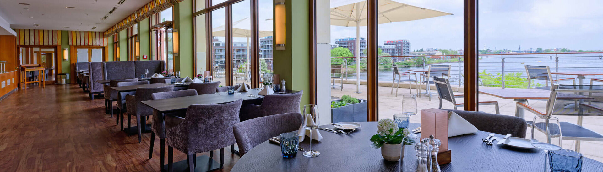 Harbour View restaurant in the ATLANTIC Hotel Wilhelmshaven Elegant hotel restaurant with covered tables, large windows and a view of a terrace with a river in the background.