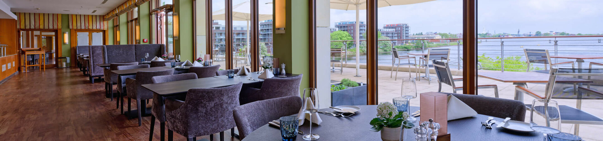 Elegant hotel restaurant with covered tables, large windows and a view of a terrace with a river in the background.