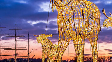 Leuchtende Elchskulpturen am Abend vor dem ATLANTIC Grand Hotel Travemünde mit Segelschiff im Hintergrund.
