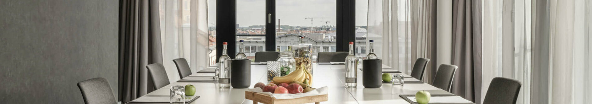 Moderner Konferenzraum im ATLANTIC Hotel Münster mit großem Tisch, Stühlen und einem Obsttablett, lichtdurchflutet durch Fenster.