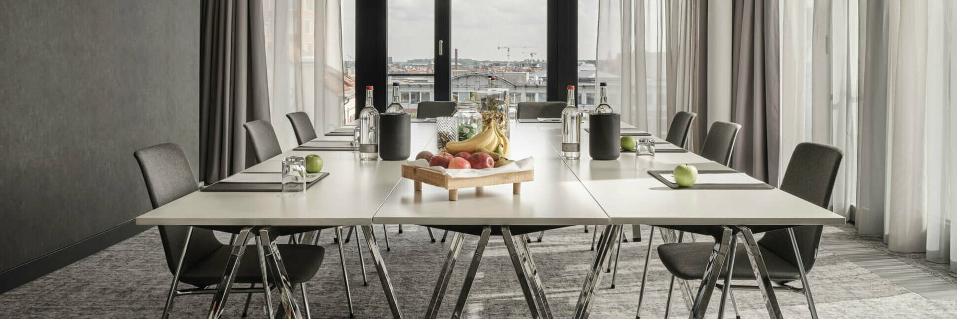 Moderner Konferenzraum im ATLANTIC Hotel Münster mit großem Tisch, Stühlen und einem Obsttablett, lichtdurchflutet durch Fenster.