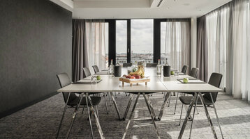 Modern conference room in the ATLANTIC Hotel Münster with a large table, chairs and a fruit tray, flooded with light through windows.