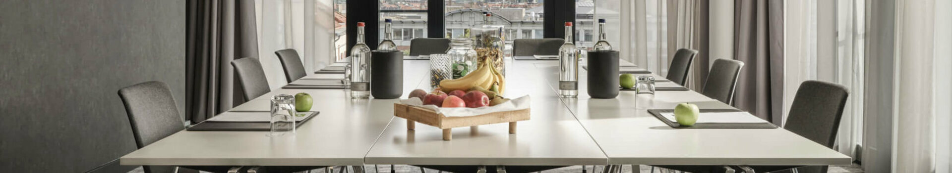 Moderner Konferenzraum im ATLANTIC Hotel Münster mit großem Tisch, Stühlen und einem Obsttablett, lichtdurchflutet durch Fenster.