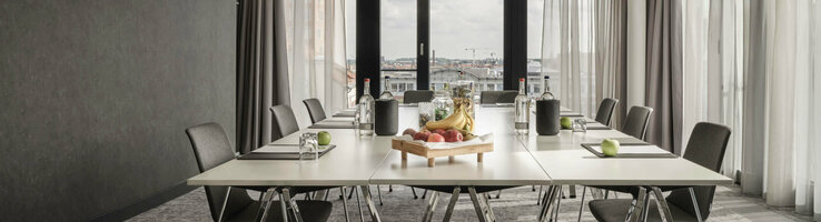ATLANTIC Hotel Münster Tagungsangebot 15 % Moderner Konferenzraum im ATLANTIC Hotel Münster mit großem Tisch, Stühlen und einem Obsttablett, lichtdurchflutet durch Fenster.
