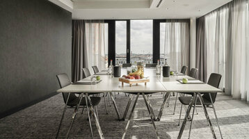 Moderner Konferenzraum im ATLANTIC Hotel Münster mit großem Tisch, Stühlen und einem Obsttablett, lichtdurchflutet durch Fenster.