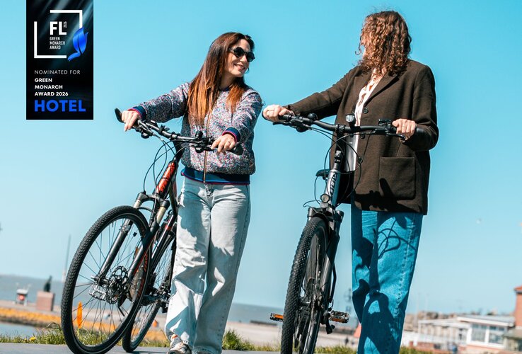 Zwei Frauen schieben ihre Fahrräder lachend an der Nordsee lang bei strahlend blauem Himmel