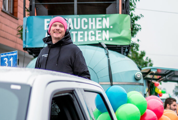 Ein lächelnder Mann mit pinker Mütze und Kopfhörern steht vor einem Wagen mit bunten Luftballons.