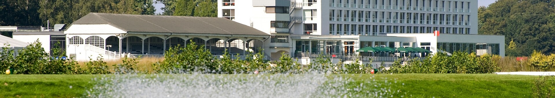 ATLANTIC Hotel Galopprennbahn in Bremen, modern building on the waterfront with a fountain in the foreground, surrounded by nature.