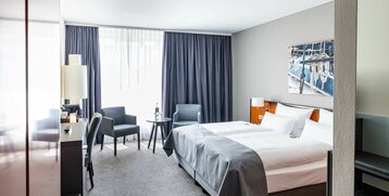 Modernes Hotelzimmer mit Doppelbett, zwei Sesseln, Tisch, Schreibtisch und großem Fenster mit blauen Vorhängen.