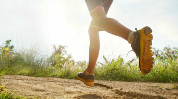 Person joggt auf einem sonnigen, grasbewachsenen Pfad unter blauem Himmel mit Wolken.