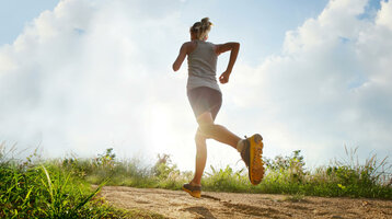 Joggen in Wilhelmshaven | Joggingkarte vom ATLANTIC Hotel Wilhelmshaven Person joggt auf einem sonnigen, grasbewachsenen Pfad unter blauem Himmel mit Wolken.