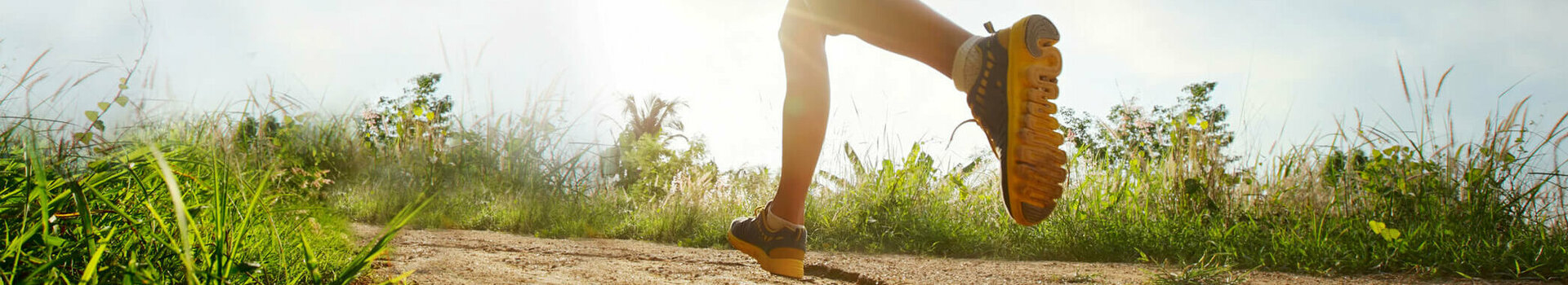 Person joggt auf einem sonnigen, grasbewachsenen Pfad unter blauem Himmel mit Wolken.