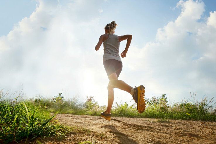 Person joggt auf einem sonnigen, grasbewachsenen Pfad unter blauem Himmel mit Wolken.
