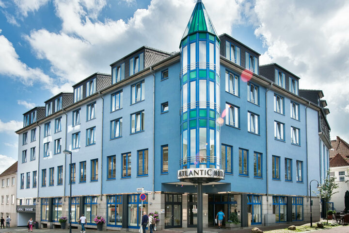 Außenansicht des ATLANTIC Hotel Vegesack - direkt im Herzen Vegesacks, Bremens wohl maritimstem Stadtteil Blaues Hotelgebäude mit markantem, verglastem Turm und Eingangsbereich, umgeben von Fußgängern unter einem bewölkten Himmel.