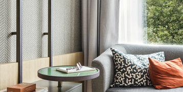 Moderne Hotelsuite mit grauem Sofa, bunten Kissen, Beistelltisch und Fenster mit Blick ins Grüne.