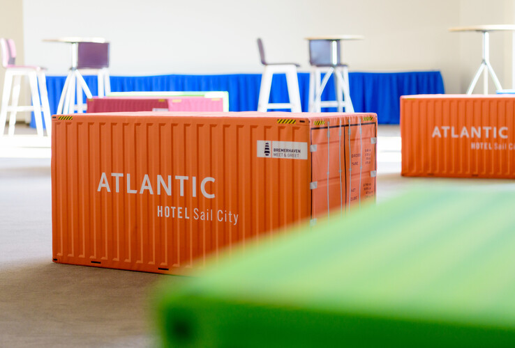 Orange Container mit "ATLANTIC Hotel Sail City" in modernem Meetingraum mit bunten Möbeln im Hintergrund.