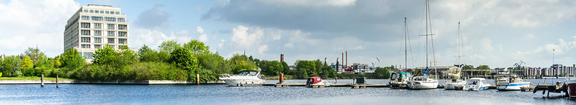 ATLANTIC Hotel Wilhelmshaven - Aussenansicht Blick auf das ATLANTIC Hotel Wilhelmshaven am Wasser mit Yachthafen und blauem Himmel im Hintergrund.