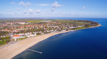 Luftaufnahme von Travemünde: Sandstrand, Promenade und Meer bei sonnigem Wetter, umgeben von grüner Landschaft und Gebäuden.