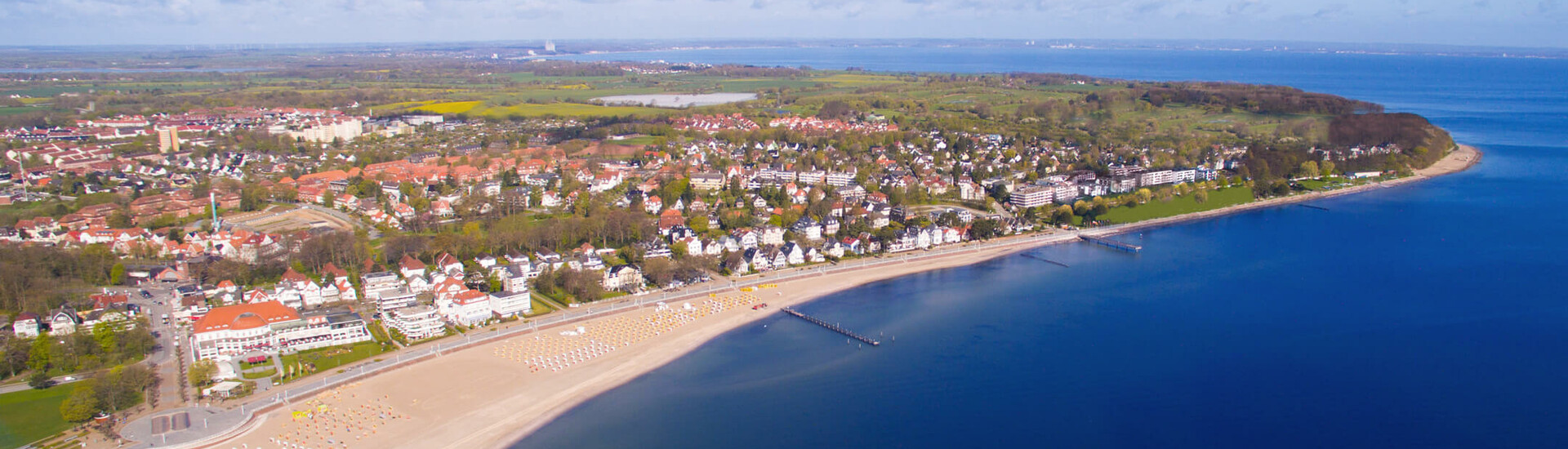Luftaufnahme von Travemünde: Sandstrand, Promenade und Meer bei sonnigem Wetter, umgeben von grüner Landschaft und Gebäuden.