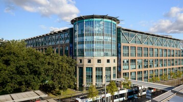 ATLANTIC Hotel Airport in Bremen Modernes Hotelgebäude mit Glasfassade, umgeben von Bäumen, mit Straßenbahn und Haltestelle im Vordergrund.