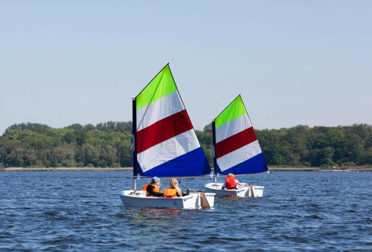Two small sailing boats with colorful sails on a calm lake, surrounded by green trees.