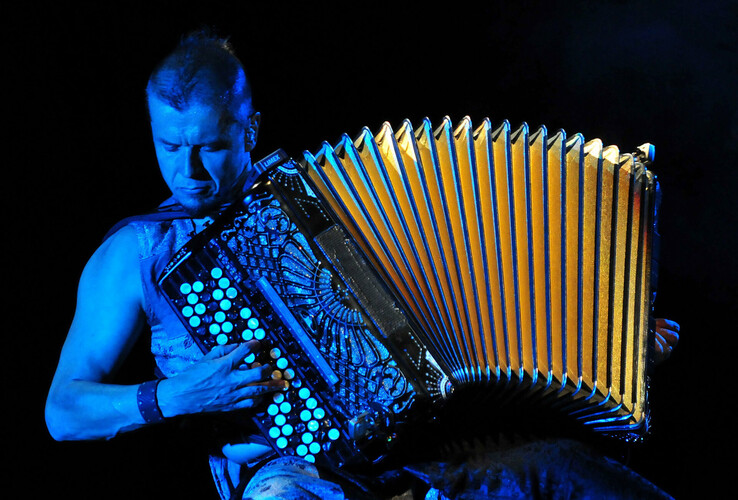 Ein Musiker spielt ein Akkordeon auf einer Bühne, beleuchtet von blauem Licht.