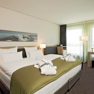 Familienzimmer im ATLANTIC Hotel Kiel Modernes Hotelzimmer mit Doppelbett, grüner Tagesdecke, Schreibtisch, großen Fenstern und stilvollem Dekor.