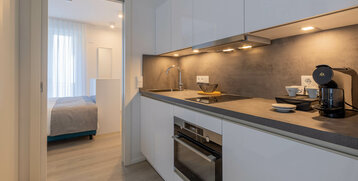 Modern hotel kitchen with white furnishings, stove, coffee machine; view into the adjoining bedroom with bed.