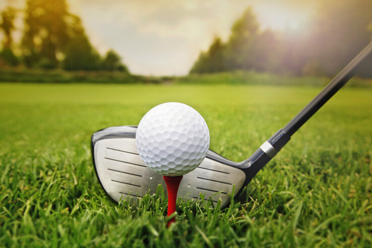 Golfball auf rotem Tee, bereit zum Abschlag, mit Golfschläger auf grünem Rasen im Sonnenlicht.