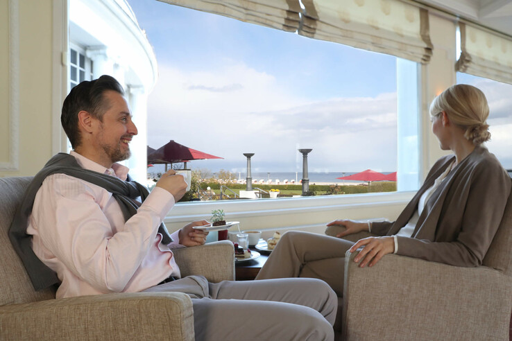 Paar genießt Kaffee im ATLANTIC Grand Hotel Travemünde mit Blick auf die Ostsee und Strandkörbe.