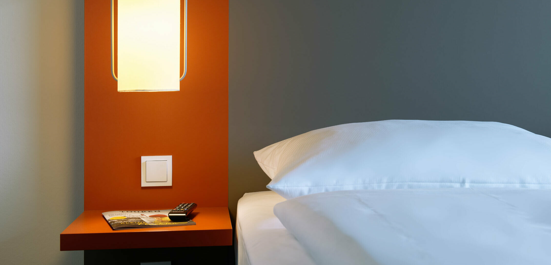 Modernes Hotelzimmer mit beleuchteter Nachttischlampe, orangener Wand, Zeitschrift und Fernbedienung auf dem Tisch.