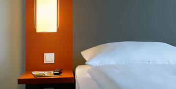 Modern hotel room with illuminated bedside lamp, orange wall, magazine and remote control on the table.