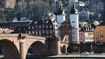 Historische Brücke mit Toren und Altstadtgebäuden im Hintergrund, umgeben von bewaldeten Hügeln.