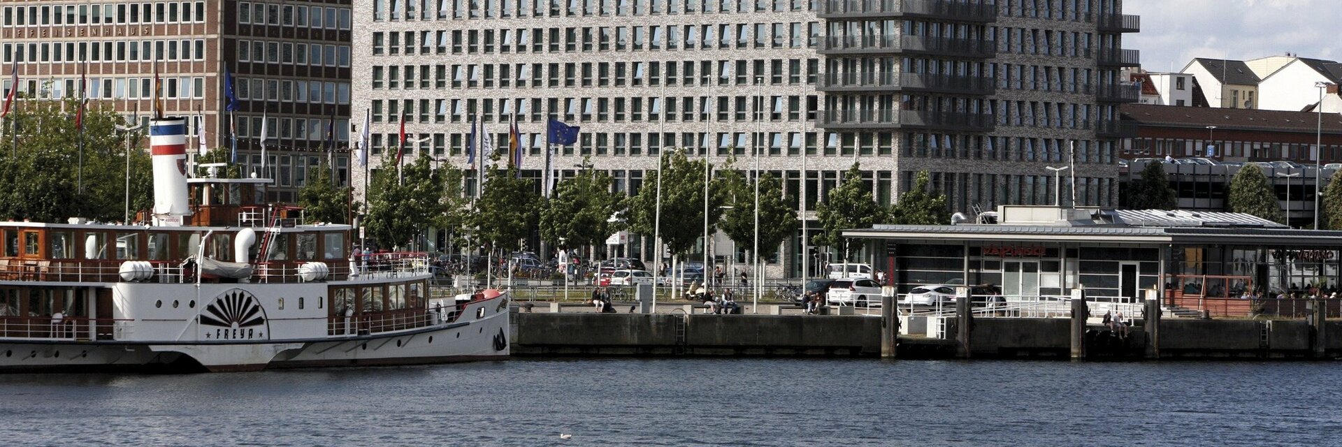 Hotel am Wasser mit modernem Gebäude, Boot im Vordergrund und mehreren Flaggen entlang der Uferpromenade.