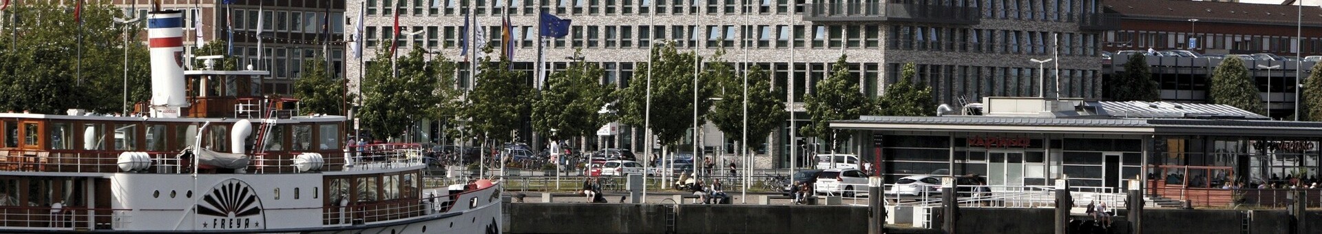 Exterior view of the ATLANTIC Hotel Kiel from across the river Hotel on the waterfront with modern building, boat in the foreground and several flags along the promenade.